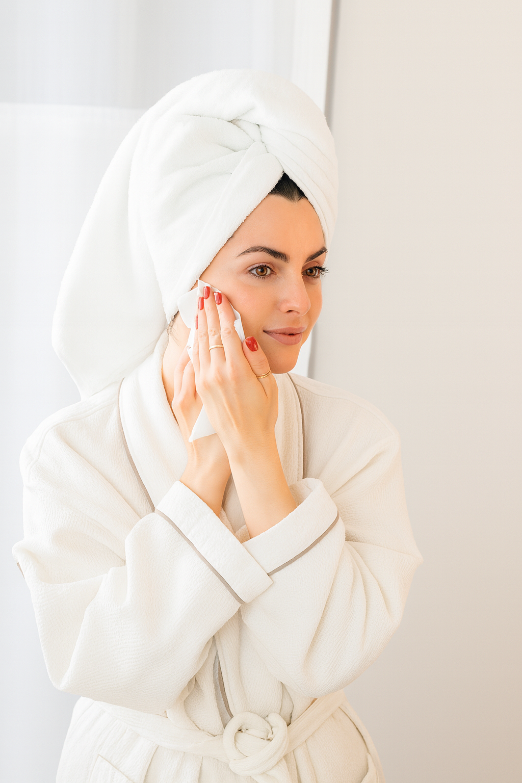 Woman in a white robe with a tissue wrapped around her head, looking at herself in the mirror.